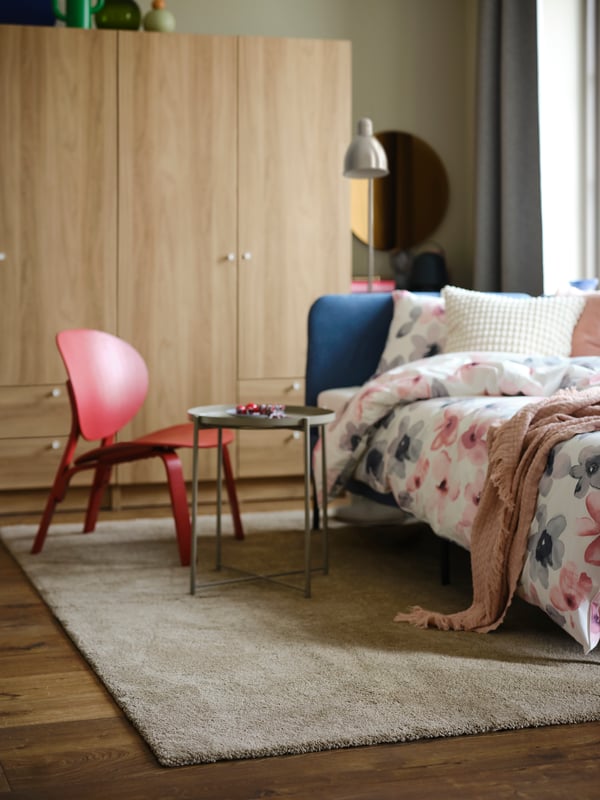 A red FRÖSET chair on top of two STOENSE low pile rugs in a bright room beside a blue BLÅKULLEN bed with corner headboard.