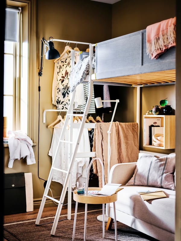 A VITVAL loft bed in a beige bedroom with a sofa and BURVIK side table underneath, and rails holding clothes to one side.
