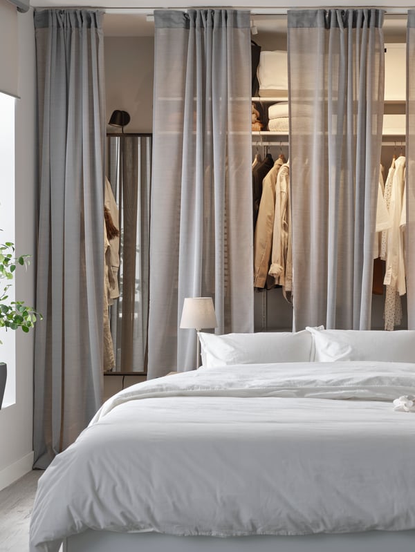 Dressing ouvert avec étagères et vétements suspendus, séparé de la chambre par des rideaux gris. Un lit blanc avec linge de lit blanc se trouve à l'avant-plan.