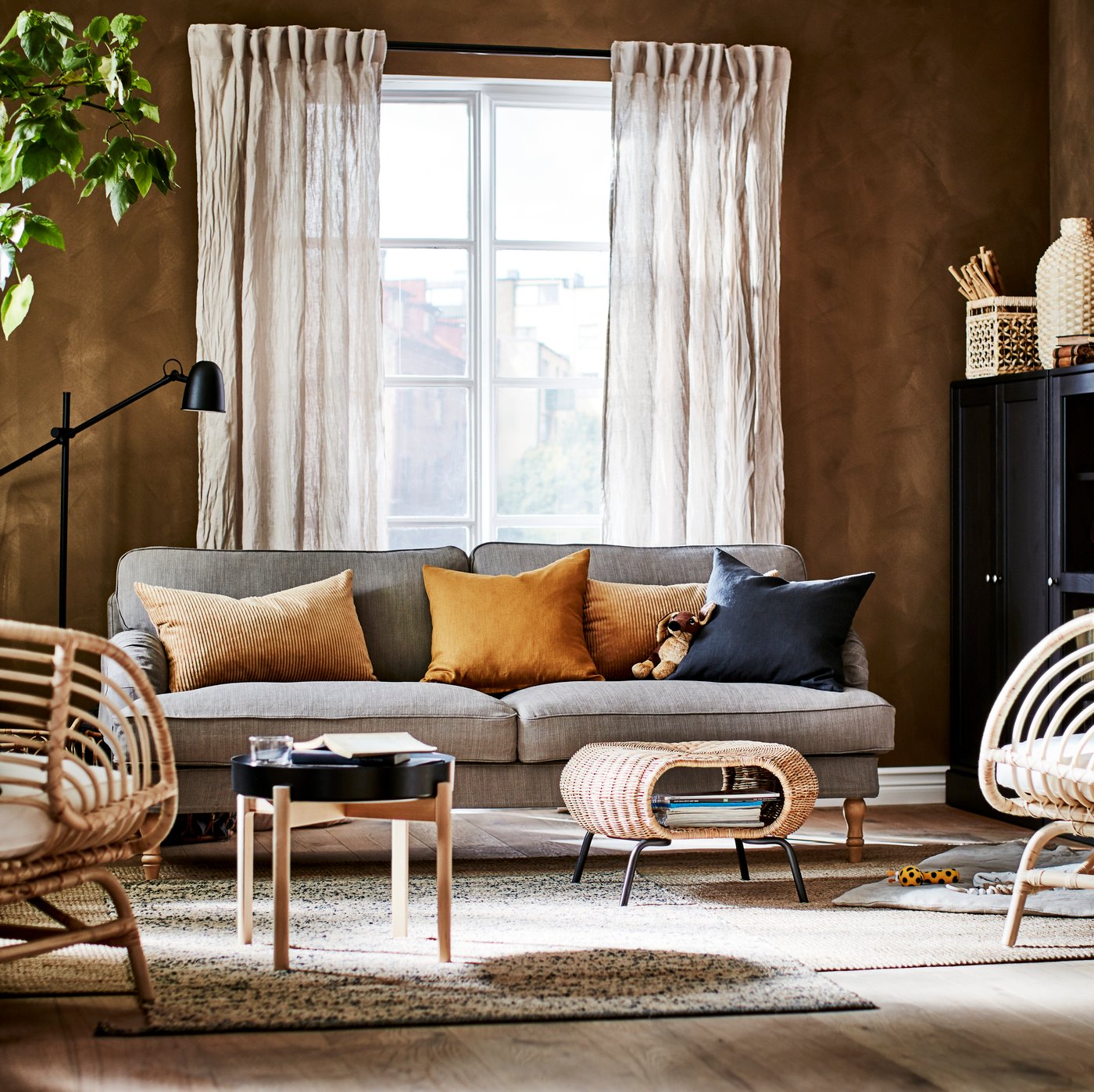 A living room with a sofa, two jute rugs, two rattan armchairs, beige linen curtains and cabinets.