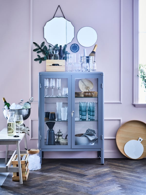 Una color gris con puertas de vidrio que permien ver los vasos y vajillas en el interior. Sobre la repisa hay unos espejos decorativos colgados en la pared.