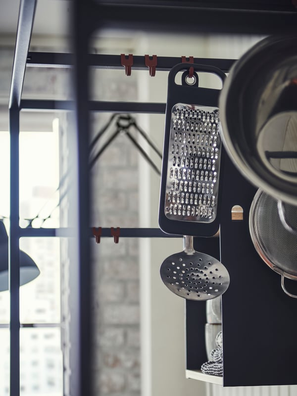 An ÄSPINGE kitchenette with an IKEA 365+ grater in black hanging along with a KLOCKREN colander and other kitchen items.