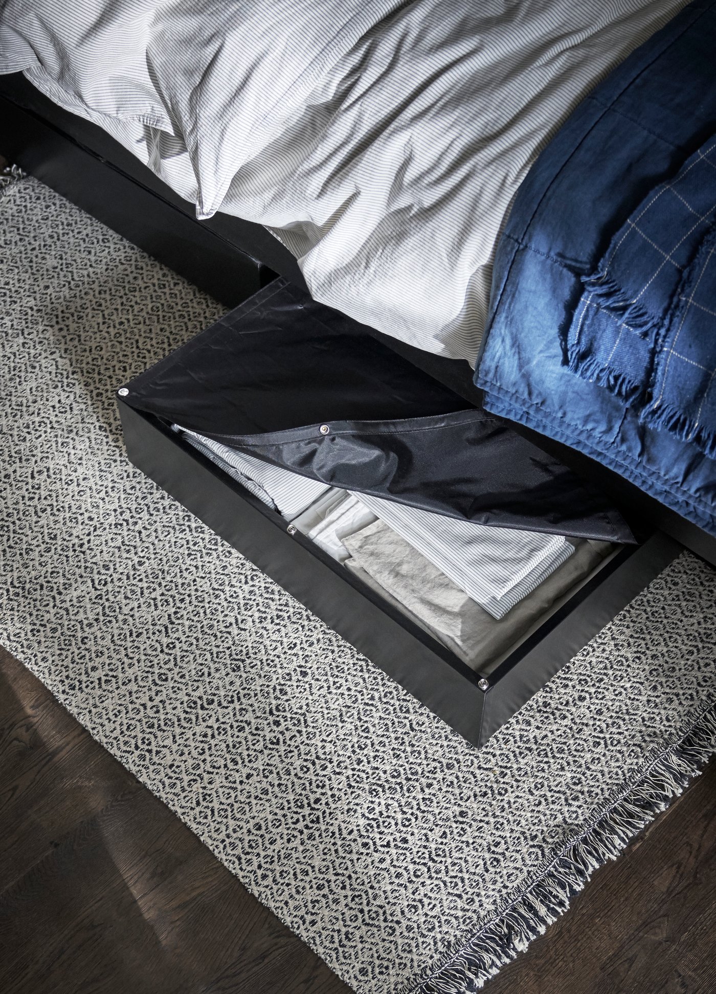 Un tiroir ouvert sous un lit aux draps blancs et couverture bleue révèle des vêtements soigneusement pliés, dont une chemise blanche, à côté d’un tapis gris à motifs.