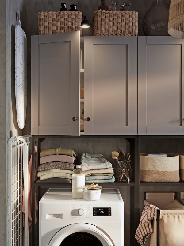 An ENHET storage combination with grey doors and open shelves stores folded clothes and baskets above a washing machine.