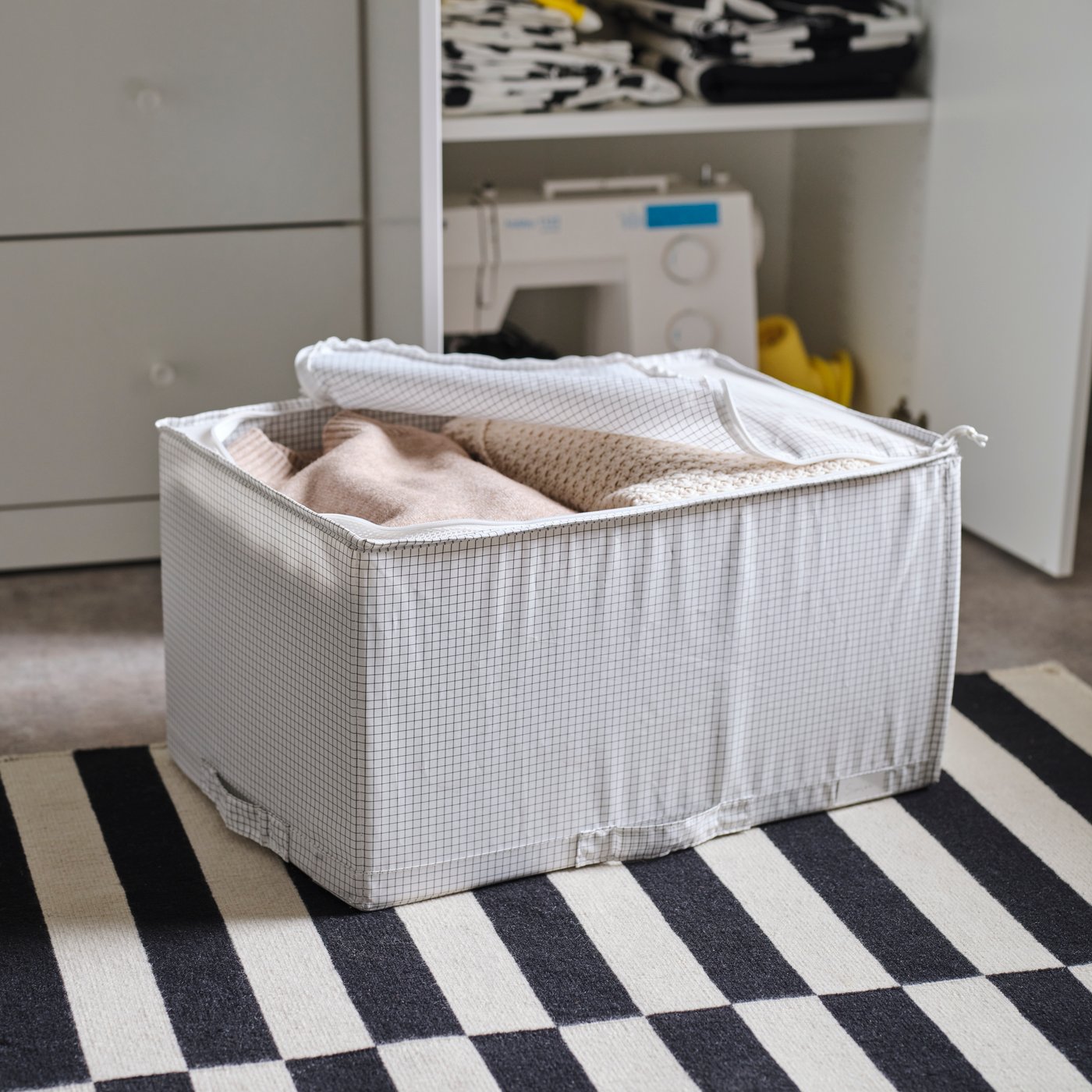 A STUK storage case containing folded clothes sits on the floor of a bedroom on a black and white striped rug.