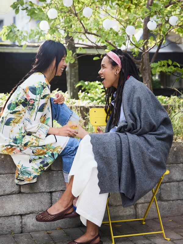 Two friends chat and laugh in a courtyard. One sits on a yellow SUNDSÖ chair, wrapped in a grey DAGGKÅPOR throw.