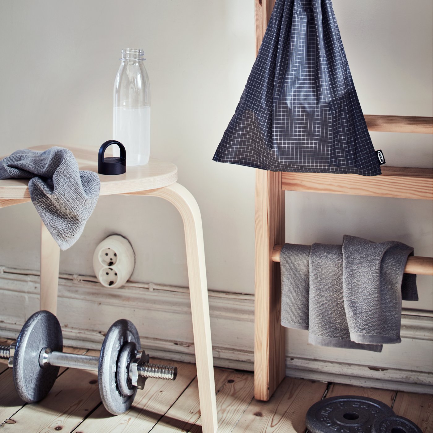 På en skammel står en vandflaske, foran ligger en håndvægt, til højre står en bænk med en grå KORNAN vaskeklud hængende henover sammen med en sort gymnastiktaske.