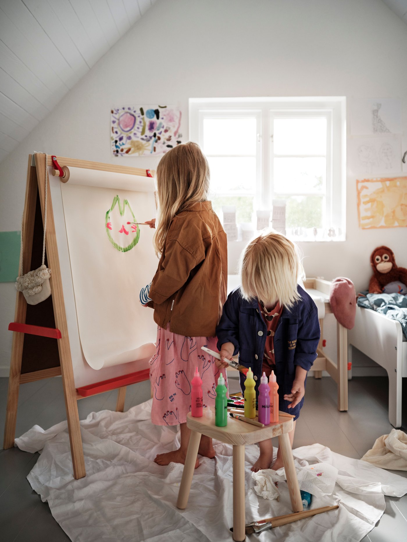 Deux enfants peignent avec créativité devant un chevalet IKEA-MÅLA, entourés de peintures colorées, de matériel de bricolage et de papier protecteur dans une salle de jeux lumineuse.