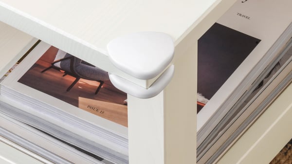 Deux protections d’angle UNDVIKA blanches ont été fixées sur le coin d’une table basse. Une pile de revues est posée sur sa surface.