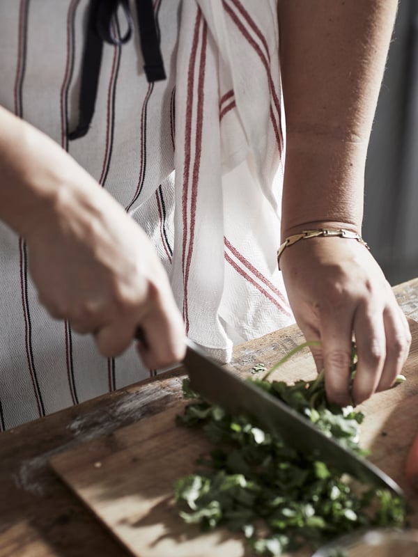 A person chopping herbs on a wooden chopping board wearing a striped apron with a red and white HILDEGUN tea towel on it.