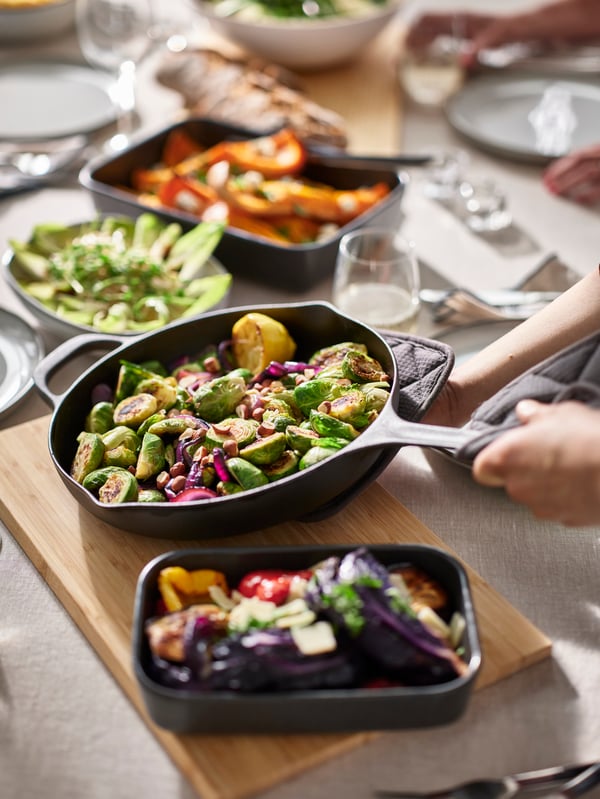 Una mesa con fuentes refractrias y un sartén VARDAGEN y otros artículos de vajilla con diferentes verduras preparadas.
