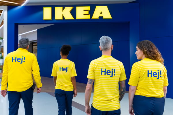 Quatre collaborateurs IKEA en t-shirts jaunes « Hej! » entrent dans un magasin IKEA, vus de dos.