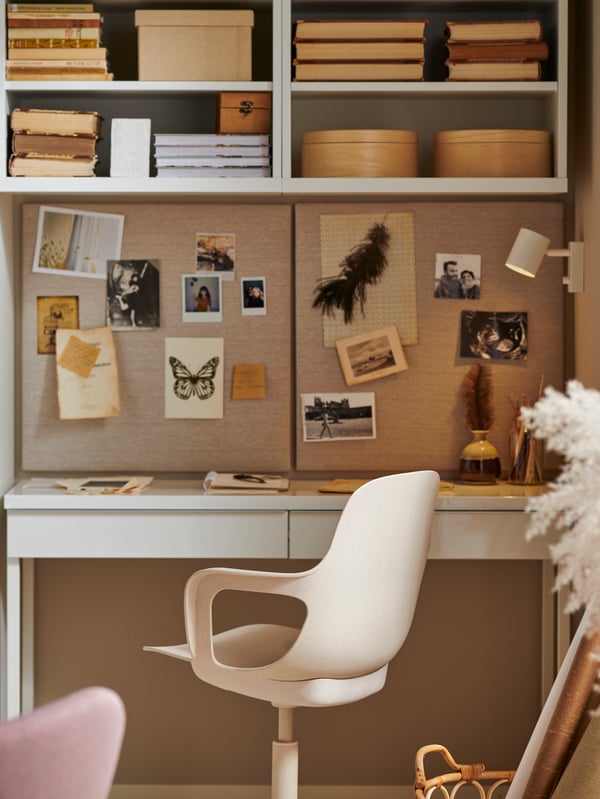Un espace de travail avec un bureau BESTÅ BURS au fini blanc très brillant, une chaise pivotante, deux tableaux d’affichage avec des punaises ainsi que des livres, des boîtes et des photos.