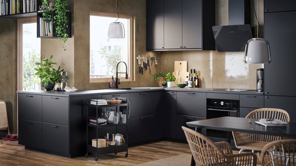 Modern kitchen setup with dark matte cabinets, open shelving with plants, hanging lights, and a small rolling kitchen cart.