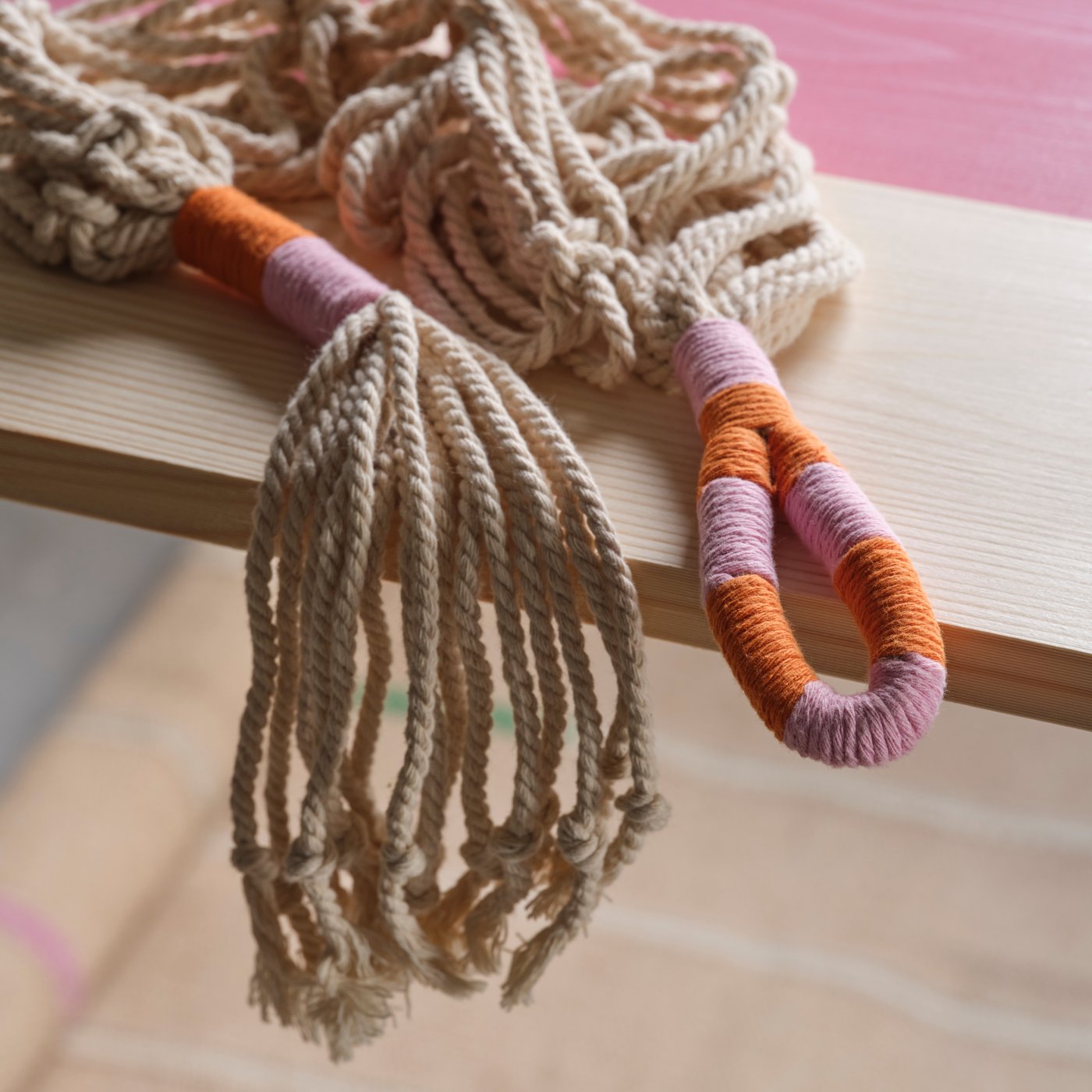 A handmade MÄVINN plant pot hanger in white macramé with details in pink and orange is resting on a wooden plank.