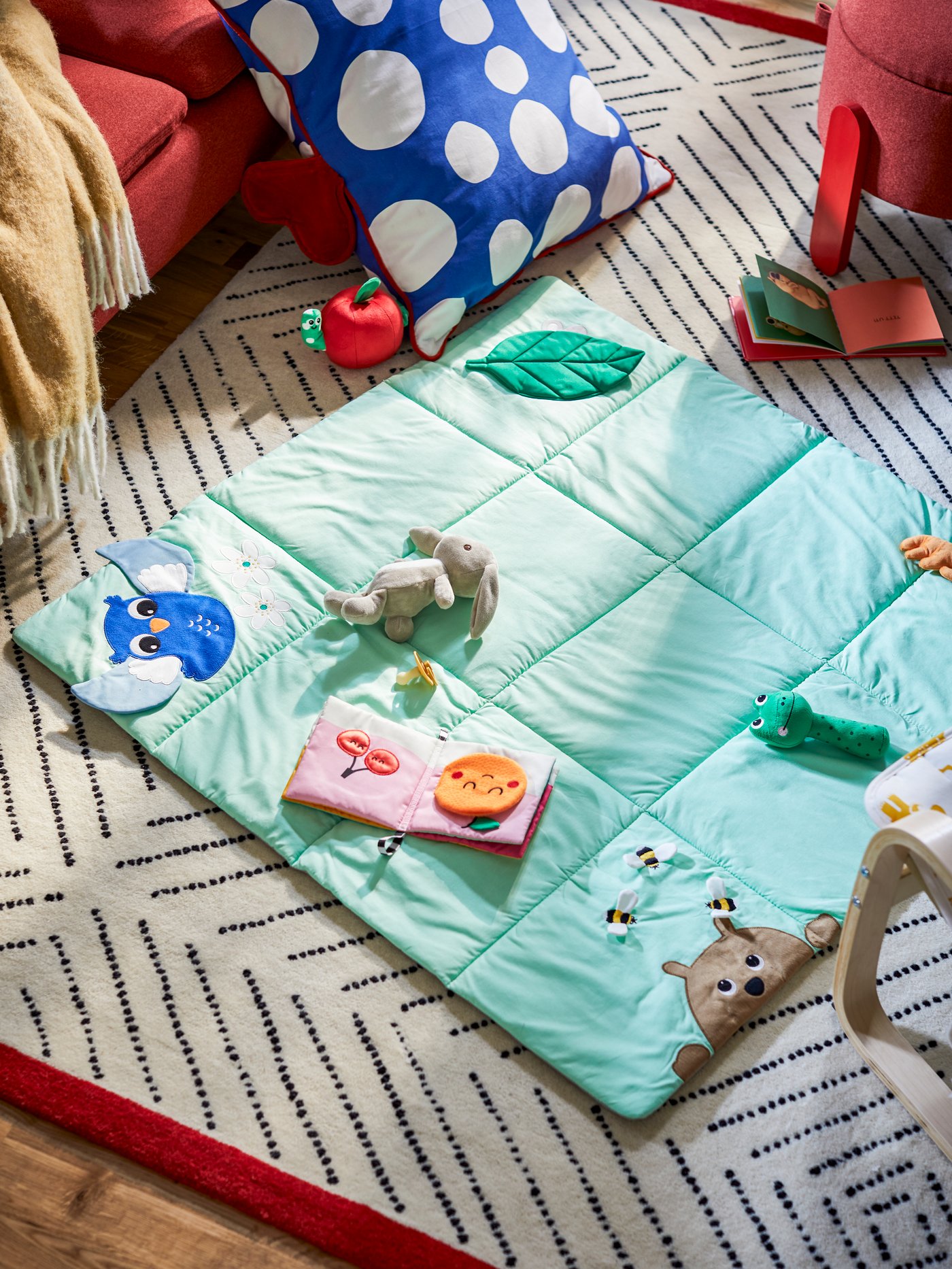 Eine SANDBI-Spielmatte und ein Kissen mit einem Bezug aus vorgeschnittenem RAKLOSTA-Stoff liegen auf einem UNDERVISNING-Teppich auf dem Boden.