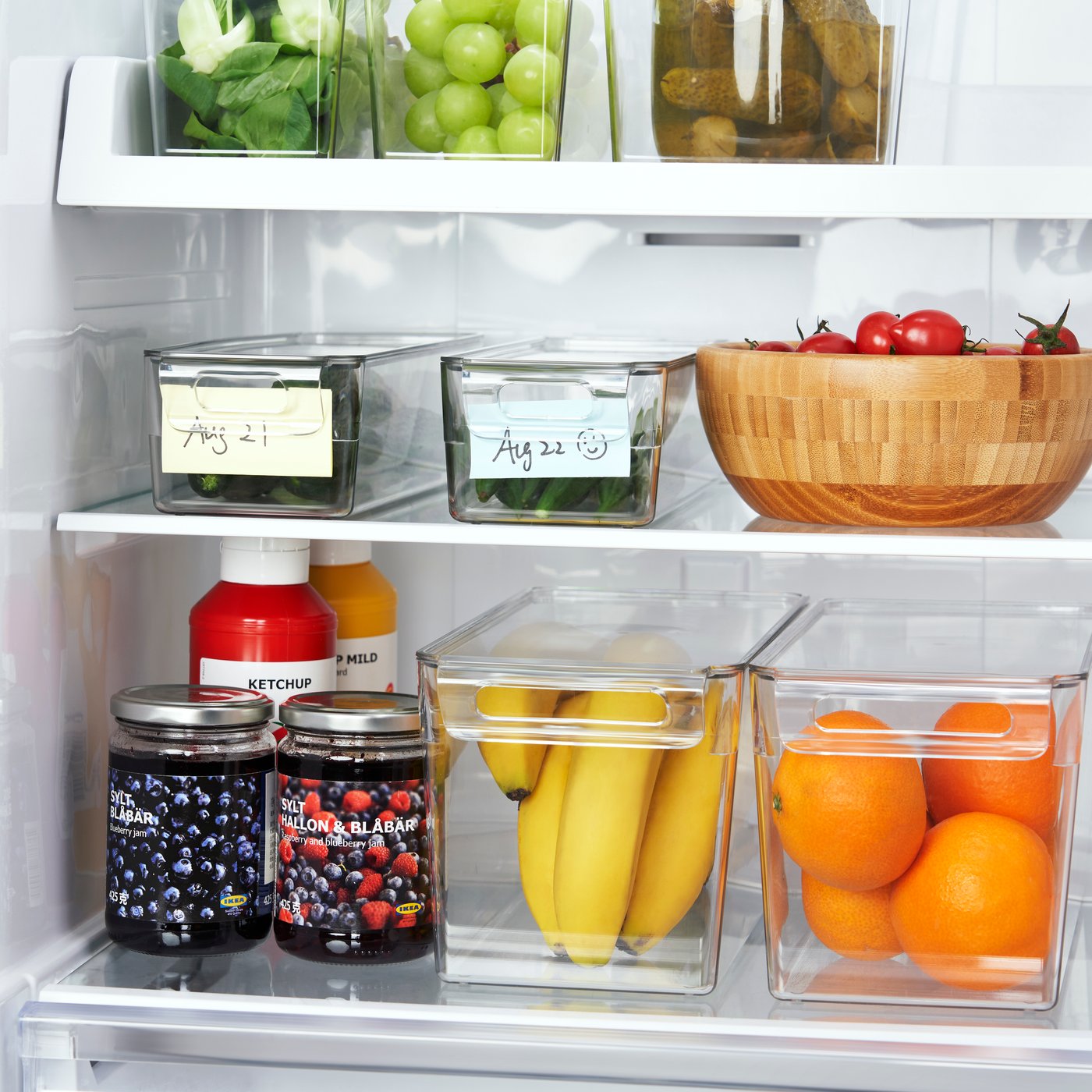 A neatly organised fridge features glass storage containers with lids, transparent bins holding bananas and oranges, jars of jam, sauces, tomatoes in a wooden bowl, and fresh produce arranged across the shelves.