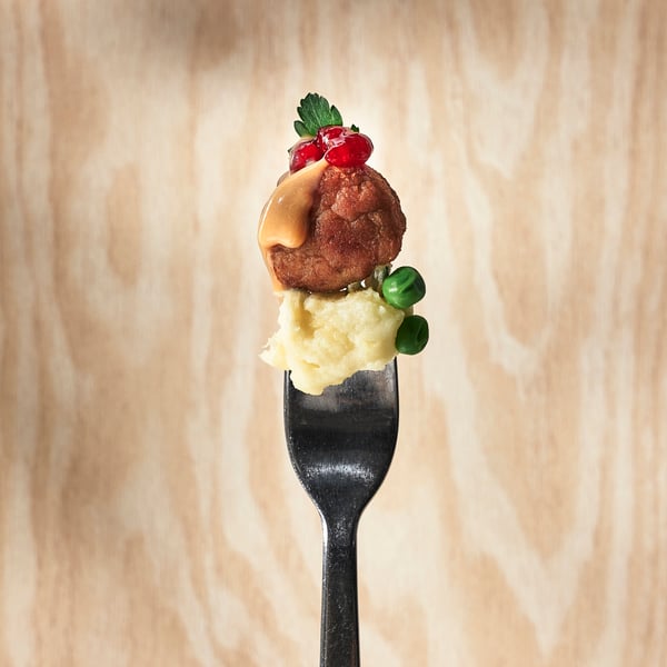 A fork holding mashed potatoes, green peas and a meatball topped with lingonberry sauce and parsley against a light wooden background.