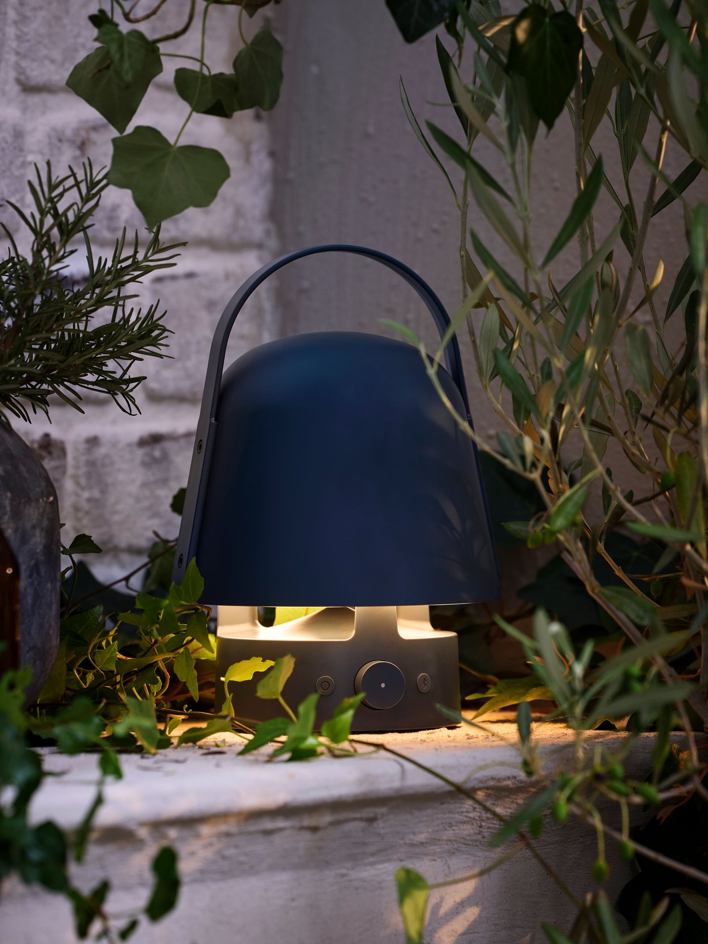 Une enceinte Bluetooth d’extérieur VAPPEBY bleue diffuse une lueur agréable près de plantes vertes disposées sur une tablette, sur une terrasse au crépuscule.