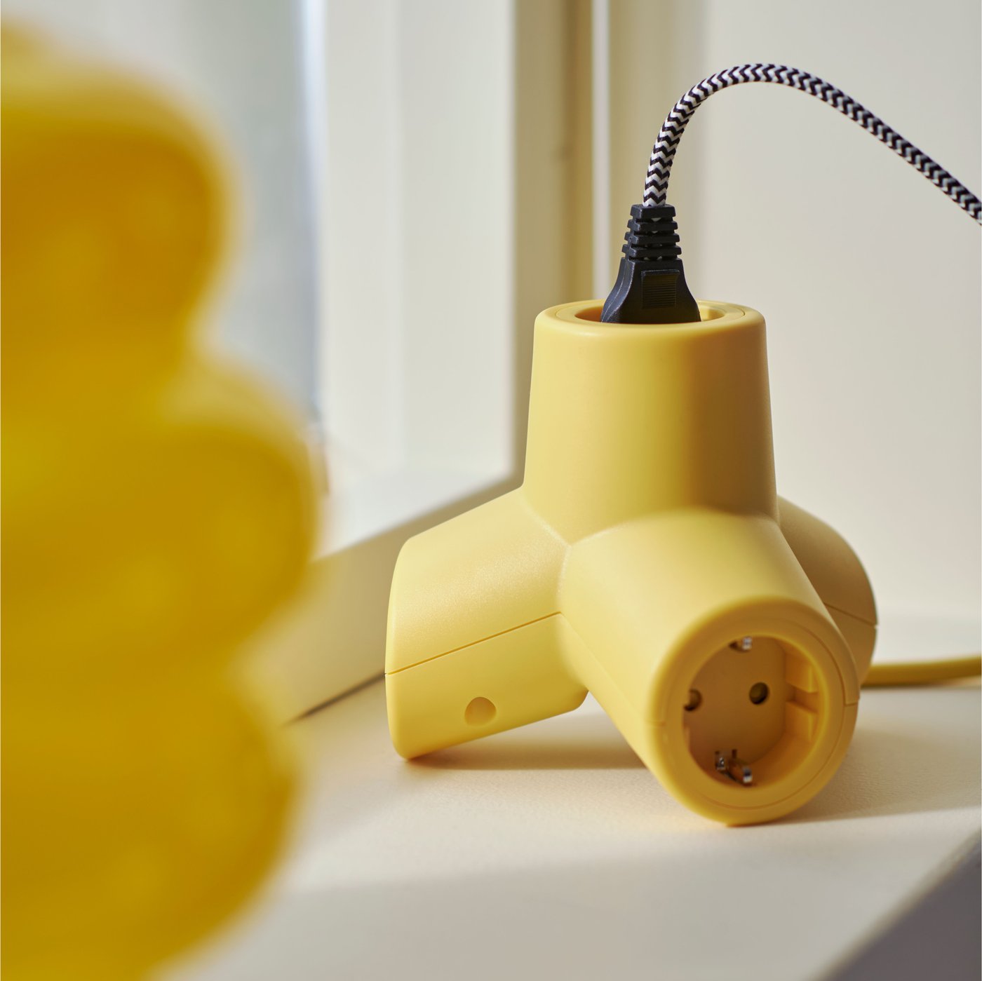 A yellow HORNFELS 4-way extension cord with one cord plugged in sitting on a window sill next to a yellow vase.