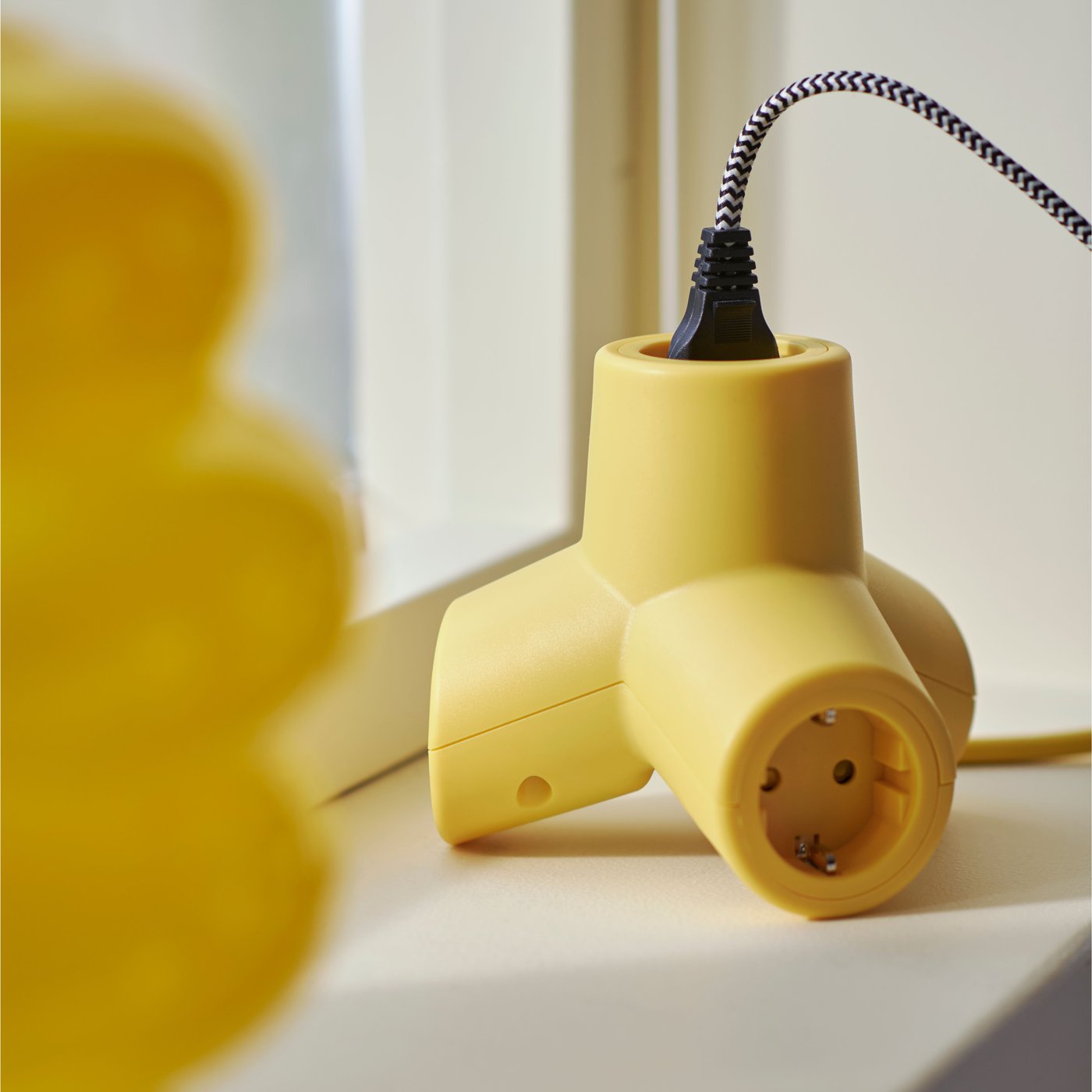 A yellow HORNFELS 4-way extension cord with one cord plugged in sitting on a window sill next to a yellow vase.