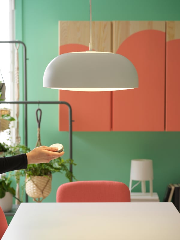 A hanging lamp in white above a white table with a chair, and a hand holding a switch with storage in the background.