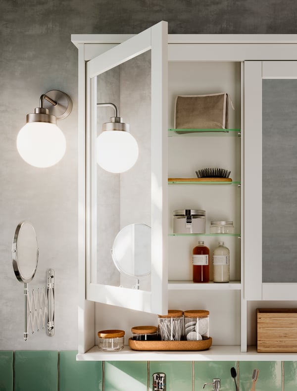 ארון עם מראת HEMNES בצבע לבן שבו אחת משתי הדלתות פתוחה מעט. לידו תלויות מראת קיר גדולה ומנורה עגולה.