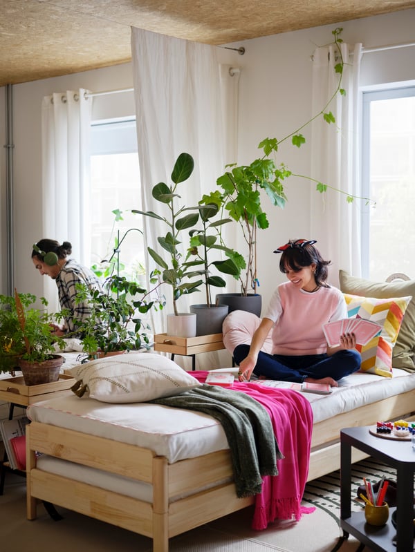 Una mujer en una cama apilable UTÅKER juega a las cartas. Detrás de una combinación de plantas y cortinas que divide la habitación, se encuentra un hombre.