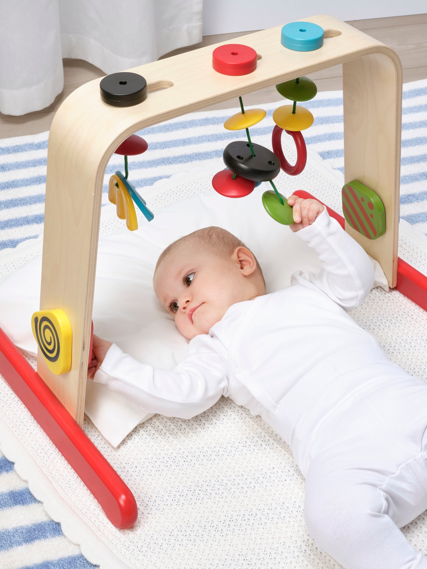 Un bébé est couché sur le dos sur une couverture tricotée blanche. Au-dessus de lui est installé un portique bébé couleur bois. On distingue derrière le pan d’un rideau.