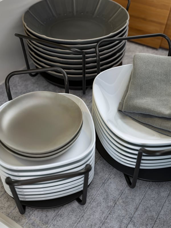 UPPDATERA plate holders in anthracite holding piles of plates and deep plates in white and grey, a grey cloth is on one.