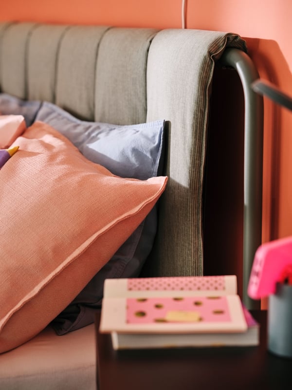 Un oreiller dans une taie ÄNGSLILJA gris bleu et un coussin dans une housse LAGERPOPPEL rose appuyés sur une tête de lit matelassée.