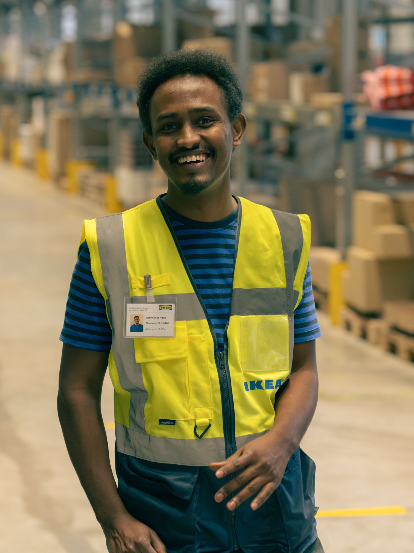 Abdi trägt eine reflektierende Warnweste und ein Hemd und geht durch ein grosses Lager voller organisierter Regale und Kartons.