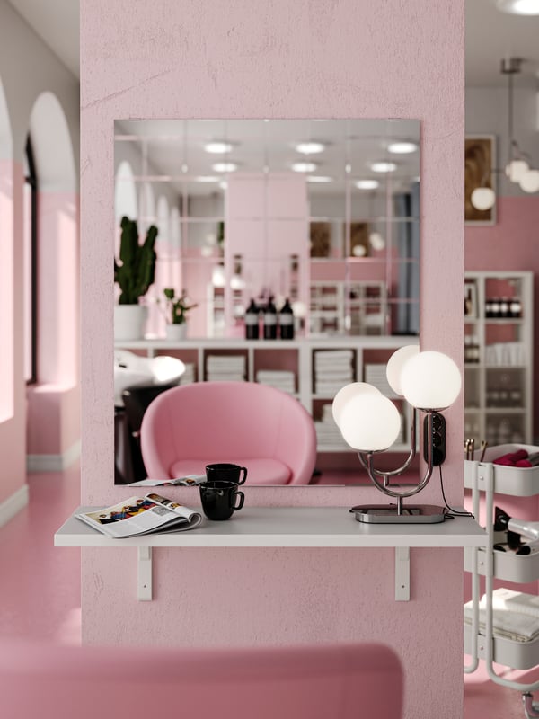 Et LETTAN spejl på en pink væg med en hylde foran danner en arbejdsstation. En kop, et magasin og en bordlampe er placeret på hylden.