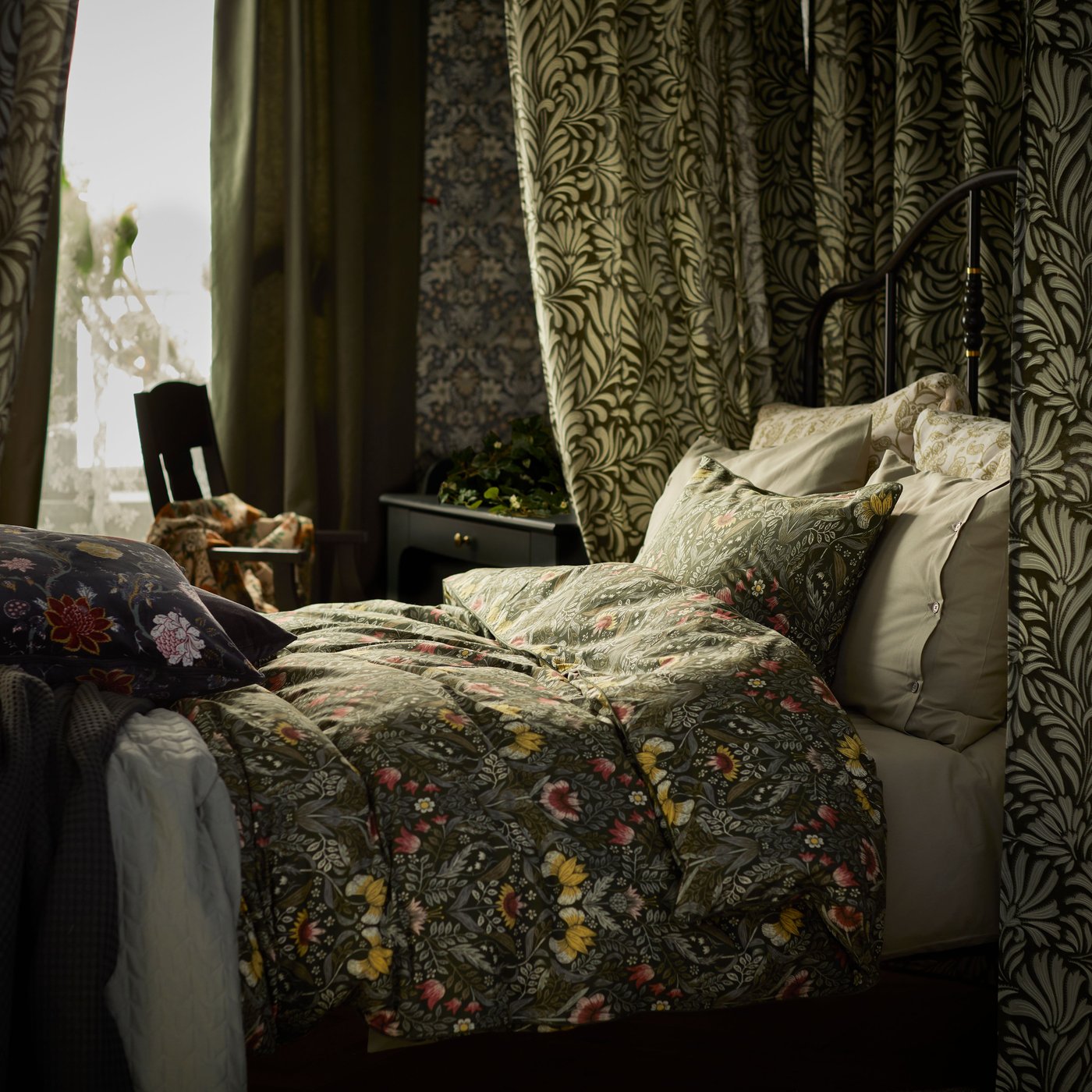 Dark green/multicolour KÄRRDUNÖRT bed linen on a bed that’s surrounded by patterned curtains, near a window.