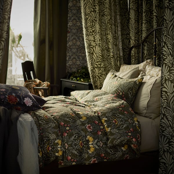 Roupa de cama KÄRRDUNÖRT em verde escuro/multicolor numa cama rodeada por cortinas com motivos, perto de janela.
