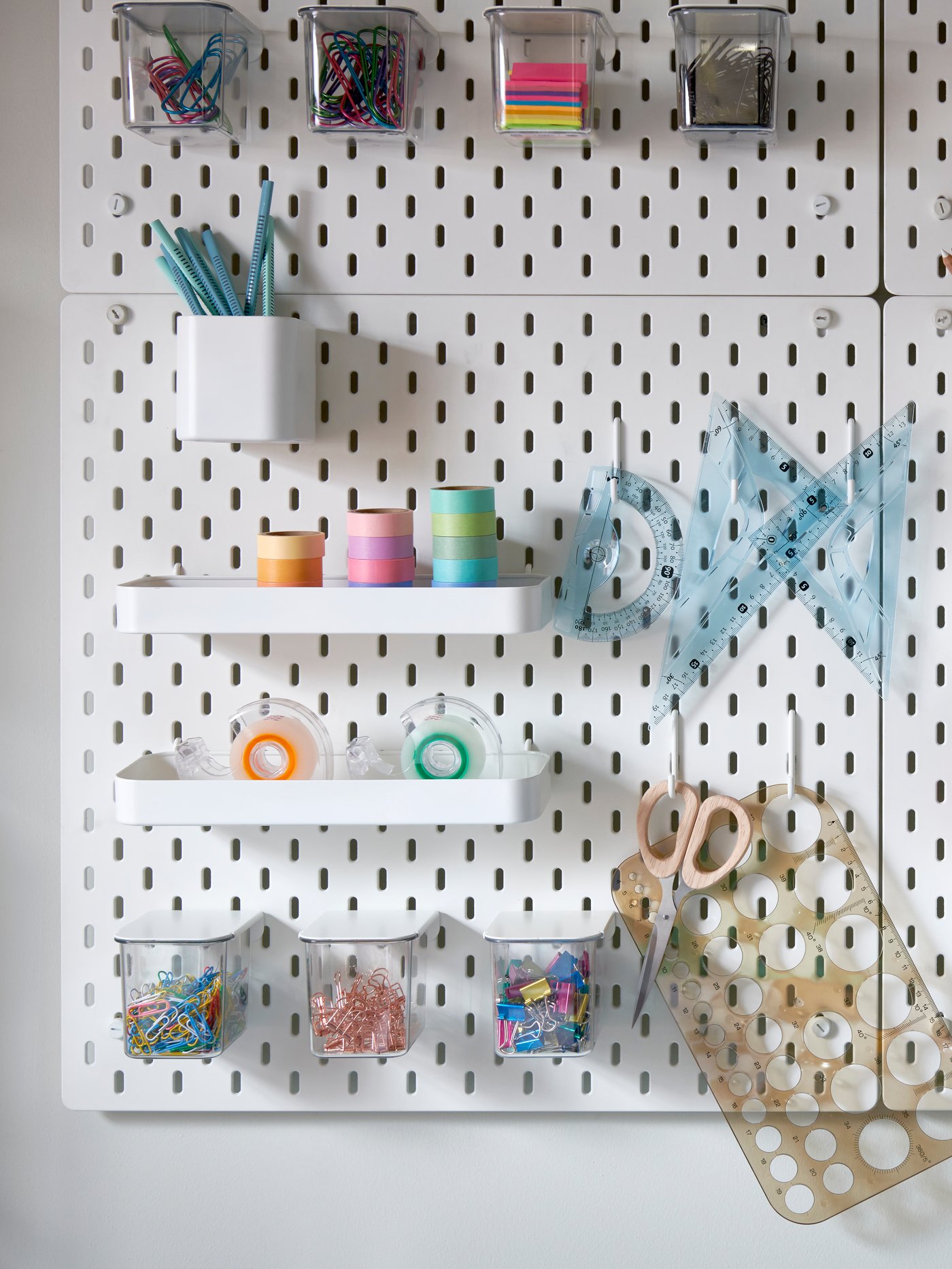A white pegboard with holes for different containers and holders storing colourful pens, paper clips, tape, rulers, etc.