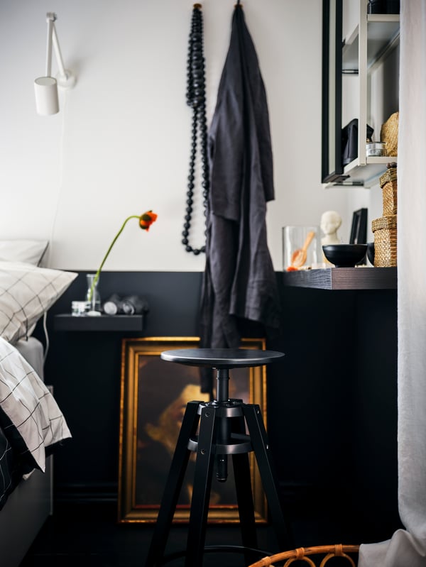 Un espace au chevet du lit avec une étagère LACK brun-noir servant de table de chevet, un tabouret de bar DALFRED noir et un meuble avec miroir ENHET.