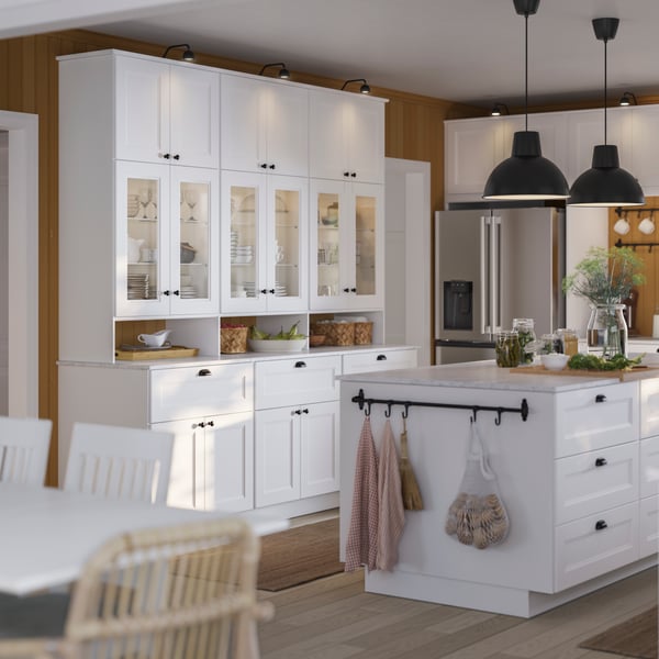 Una cocina AXSTAD blanca con una isla grande, gabinetes con frentes de vidrio y aparatos de acero inoxidable, además de iluminación colgante.