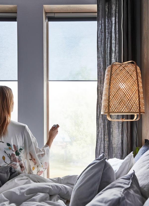Person sits on a bed, controling KADRILJ smart blinds with a remote control. A natural fibre lamp hangs above the layered textile bed. 