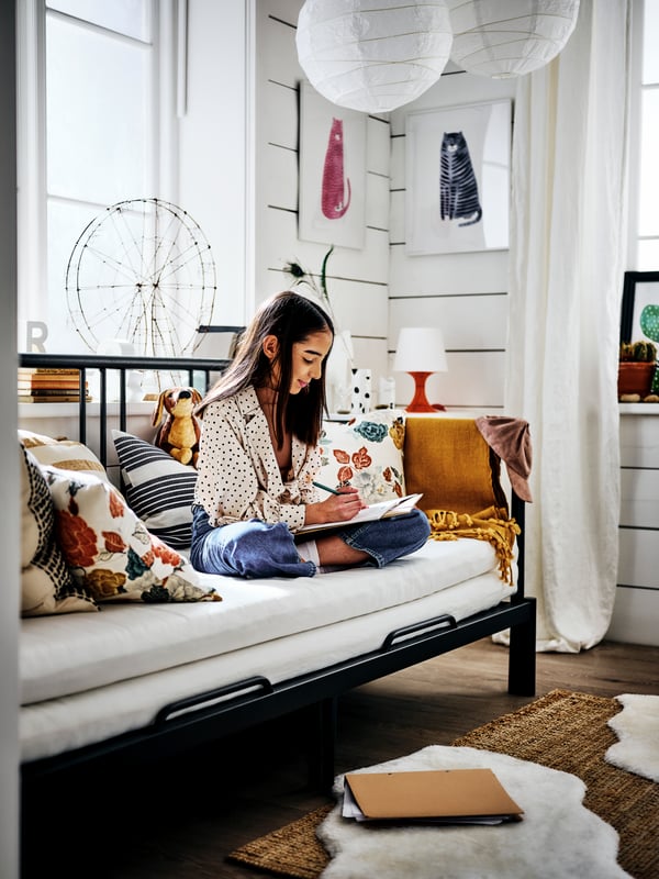 Une fille est assise sur un lit banquette avec structure métallique noire avec des coussins et un plaid jaune dessus, dans une pièce avec du lambris blanc.