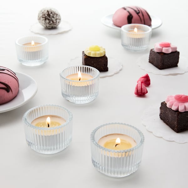 Five FINSMAK glass tealight holders with lit candles, on a white dining table next to various pastries and pieces of cake.
