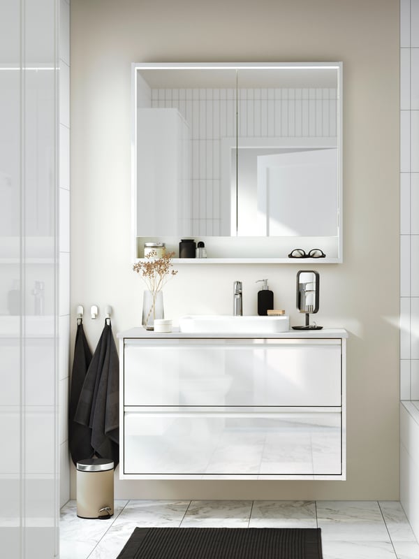 Un meuble lavabo ÄNGSJÖN doté de tiroirs en blanc très brillant avec une armoire à pharmacie au-dessus et deux serviettes sur les crochets à côté.
