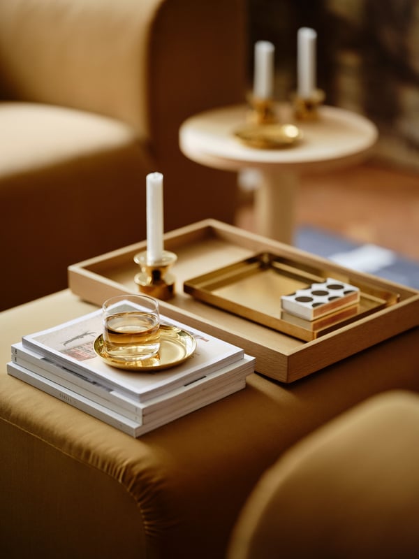 An oak veneer STOCKHOLM 2025 tray, a brass dish, tray, and candlestick/tealight holder, and more on top of a beige pouffe.