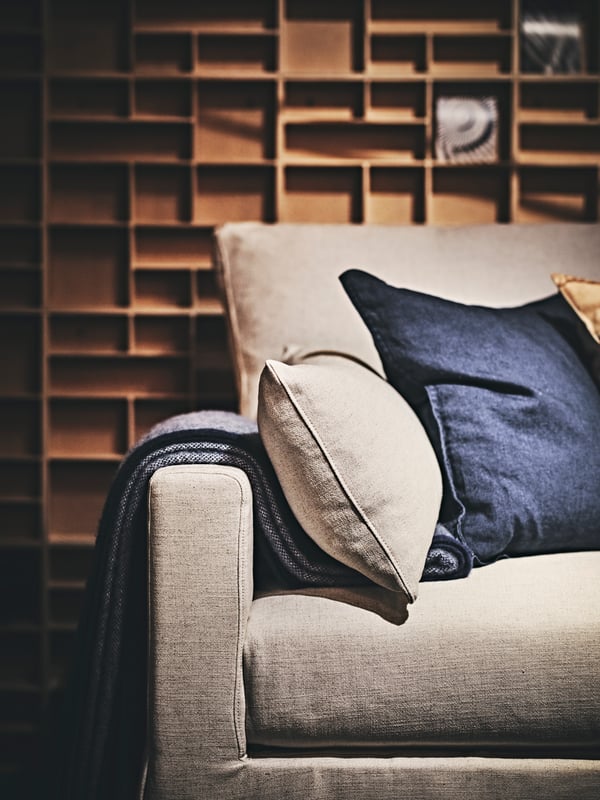 A close-up of a beige sofa with three pillows in beige, blue and yellow, and a dark blue blanket. And a wooden wall shelf.