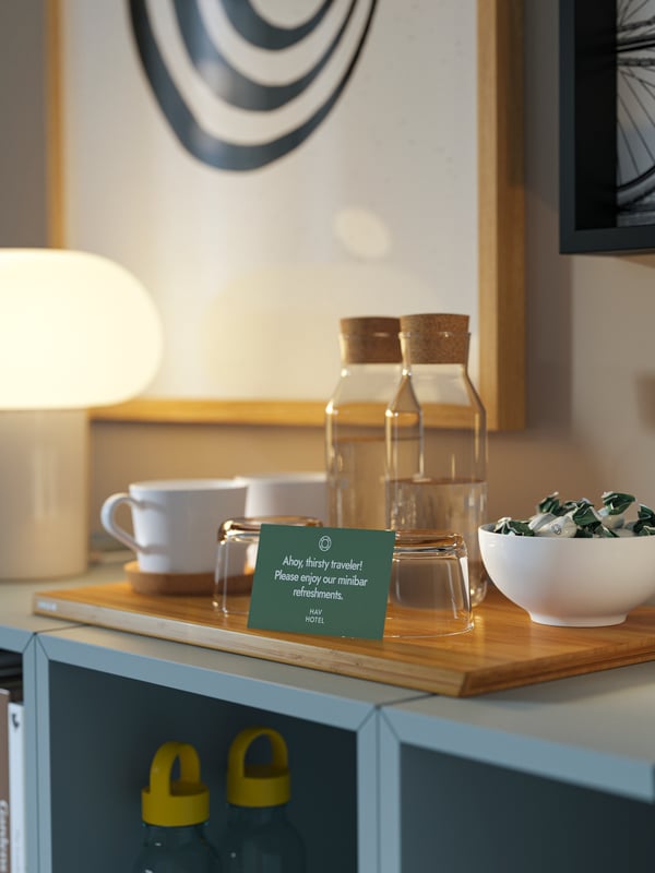 Dva IKEA 365+ karafa sa zapušačem od bistrog stakla, bele šolje, čaše i činija sa slatkišima na dasci za sečenje na EKET elementima.