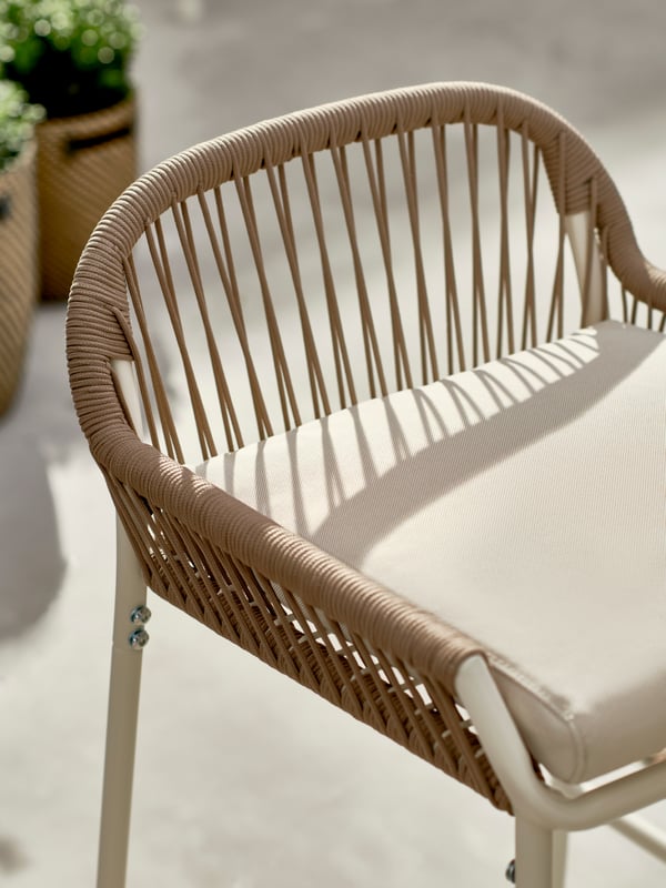 A white-and-beige SEGERÖN bar stool, placed in a light, sunlit spot with leafy potted plants on the ground beneath.