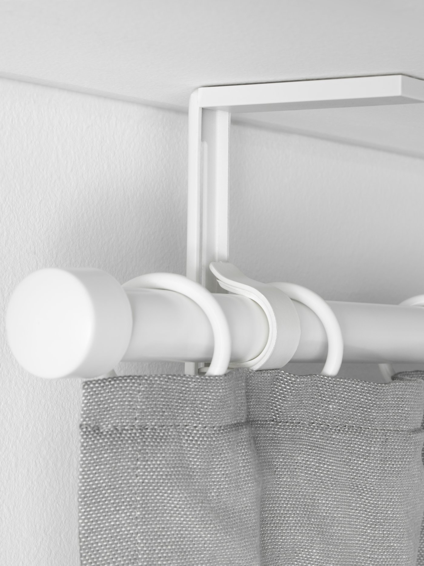 A white BETYDLIG ceiling bracket with a white RÄCKA curtain rod attached, holding a light grey curtain with rings.