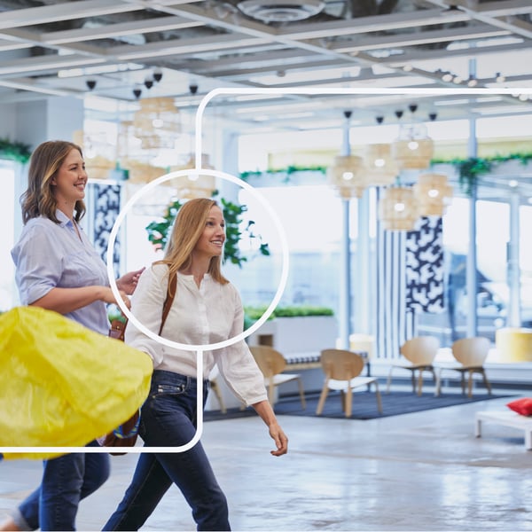 Dve žene šetaju u zatvorenom prostoru s IKEA torbama i smeškaju se. 