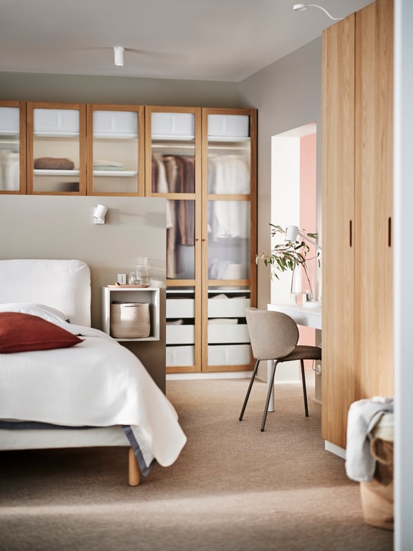 A white SAGESUND upholstered bed covered in white bed linen in front of a PAX/TONSTAD wardrobe in white and oak veneer/glass.