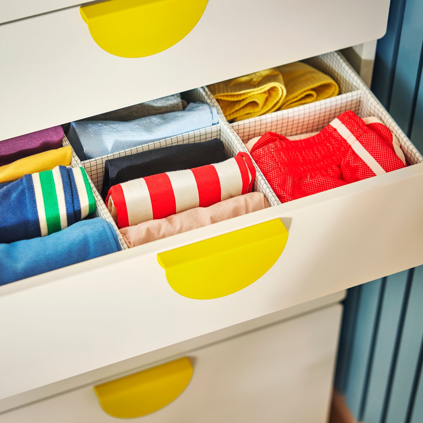 Un jaladera BEGRIPA en color amarillo en un cajón blanco, abierto para mostrar ropa infantil colorida y cuidadosamente doblada en compartimentos.